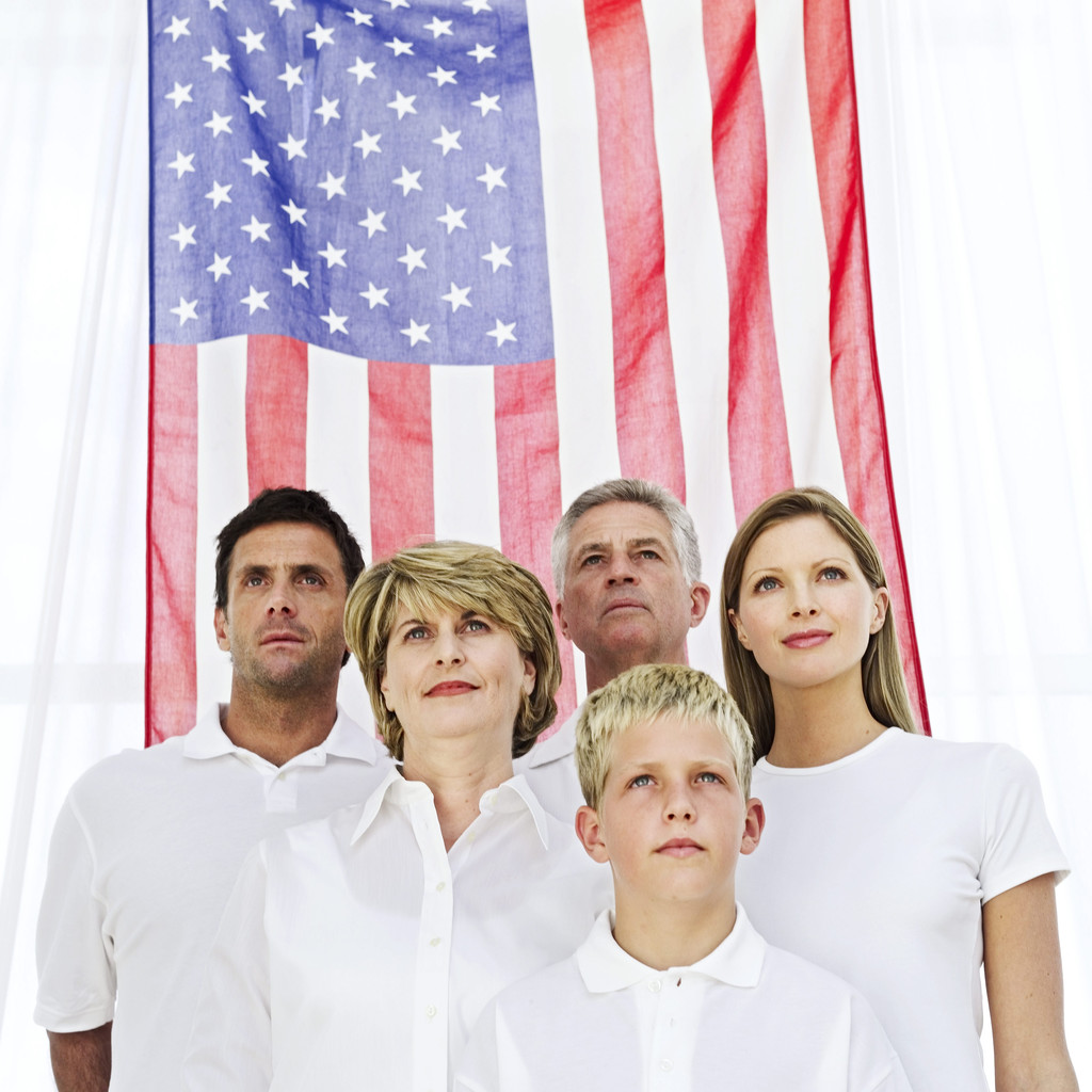 Five people are standing in front of a large American flag, looking upward. They're wearing white shirts, with a mix of men and women, and one child. The group appears to be solemn or reflective.